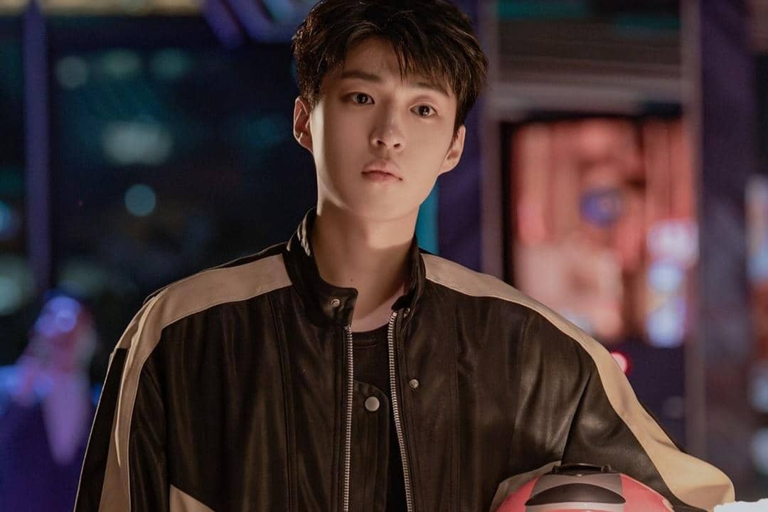 Seorang pria muda mengenakan jaket kulit hitam dengan ekspresi serius memegang helm merah di latar malam berlampu neon.